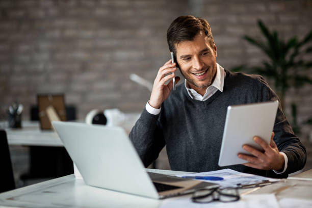 hombre de negocios feliz usando tableta digital mientras habla en el teléfono celular en la oficina. - track pad fotografías e imágenes de stock