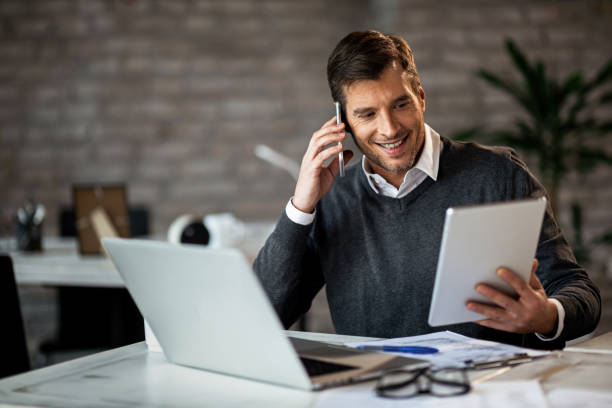 Happy businessman using digital tablet while talking on cell phone in the office.