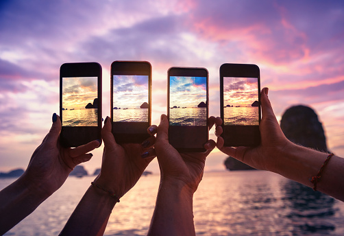 Closeup photo of four hands with mobile phones taking photo of beautiful sunset over sea bay