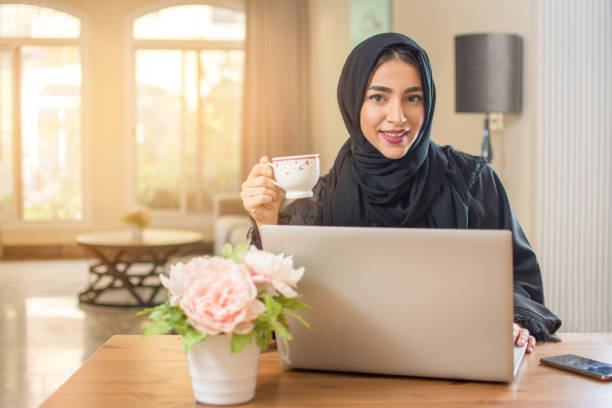 istock Young Arab woman with coffee cup using laptop and looking at camera 1132474832