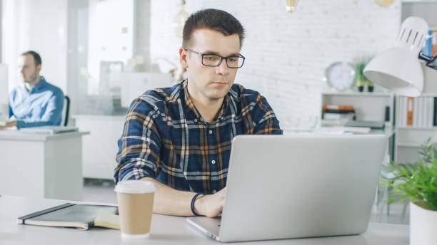 istock Face Front Man Sitting at His Creative Office Table Working on His Laptop. In the Background man Working at Their Desktops. 1132478927