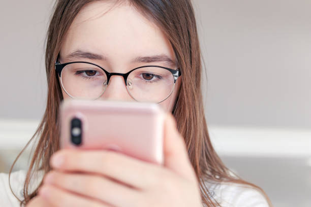 Close-up portrait of cute serious tween girl in glasses looking at smartphone in her hands. Child and gadget concept. Internet safety and social networks addiction. Vision problem. stock photo