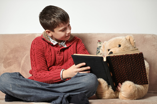 boy reads a book, sits on the sofa with a bear toy