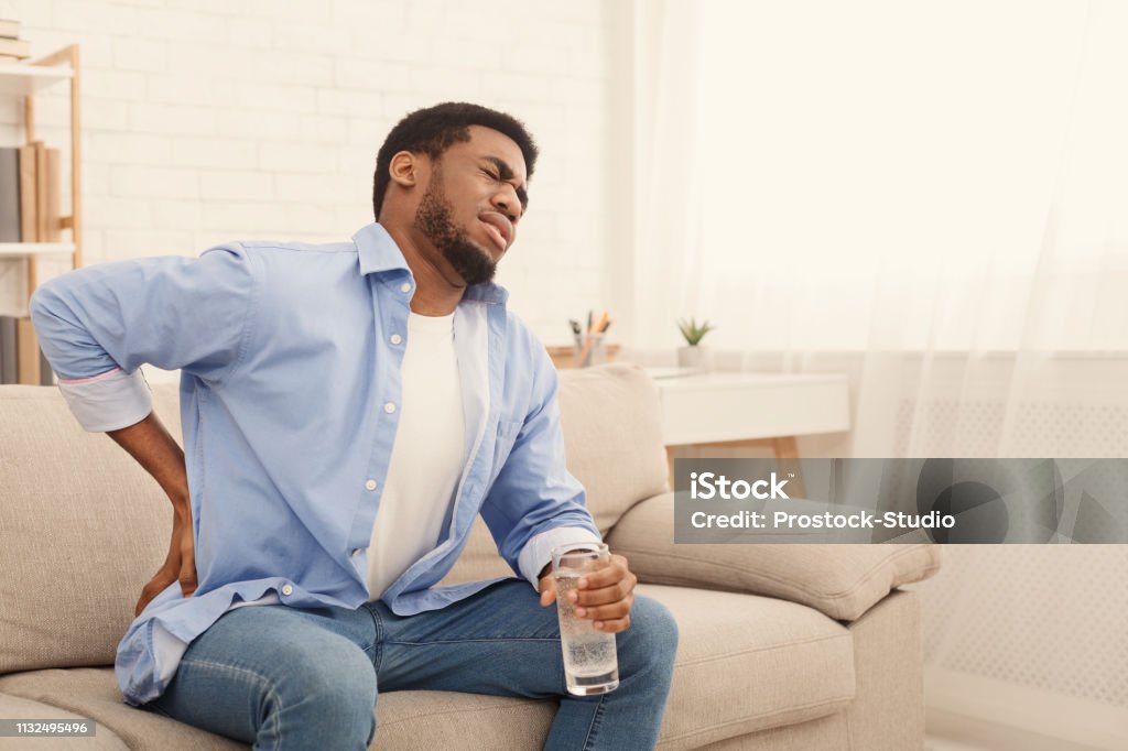 Young black man with back pain at home Young african-american man with back pain, pressing on hip with painful expression, sitting on sofa at home with glass of water, copy space Pain Stock Photo