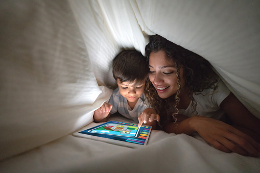 Portrait of a happy mother and son playing on a digital tablet in bed and smiling under the covers