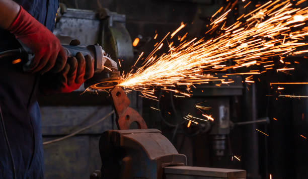 Body shot of mechanical engineer worker wearing safety gloves and operating a angle grinder on his workbench to metal held in an iron vice inside a workshop with flash sparks stock photo