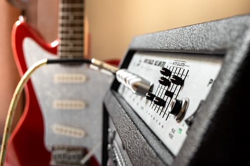 Red electric guitar plugged into large speaker. Close up of amplifier controls with blur effect on background. Playing guitar in home.