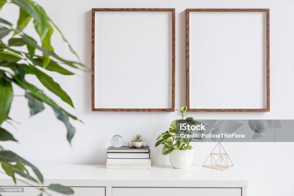 Stylish home interior with two brown wooden mock up photo frames on the white shelf with books, beautiful plants, gold pyramid and home accessories. Minimalistic concept of white room decor. Minimalistic home decor of interior with mock up frames. Modern concept of white room with plants. Picture Frame Stock Photo