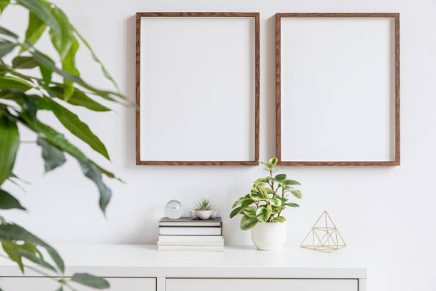 Stylish home interior with two brown wooden mock up photo frames on the white shelf with books, beautiful plants, gold pyramid and home accessories. Minimalistic concept of white room decor. Minimalistic home decor of interior with mock up frames. Modern concept of white room with plants. surrounding wall photos stock pictures, royalty-free photos & images