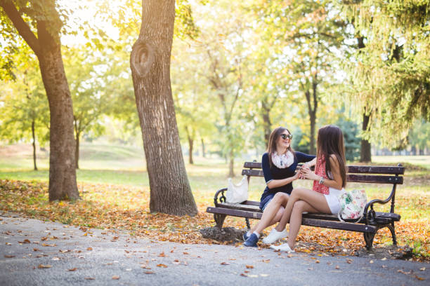 Two young beautiful caucasian girls in summer dresses sitting on a park bench and talking.