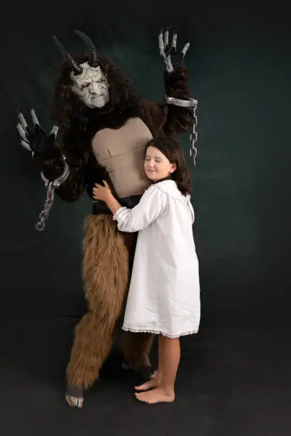 Young girl in white robe snuggles with a scared Krampus