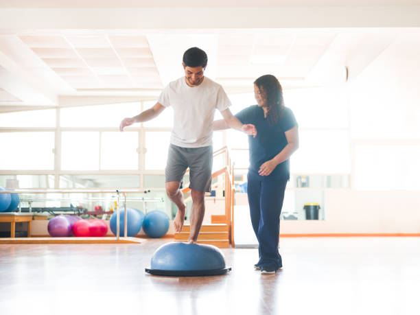 Male physical rehab patient standing on bosu ball A young latin man standing on an balance trainer with one leg next to a female occupational therapist in a physical therapy room. sports medicine photos stock pictures, royalty-free photos & images