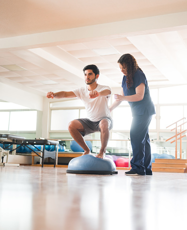 A young latin man squatting on a bosu balance ball next to a physical therapist and looking away.
