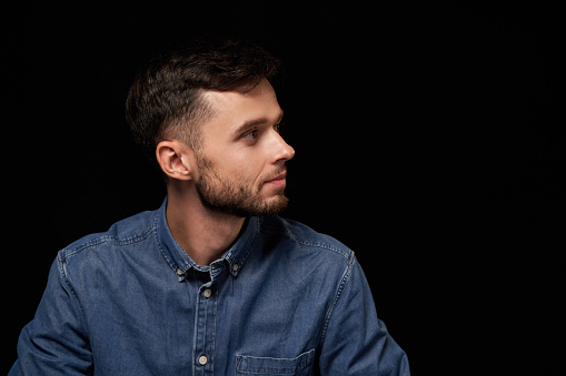 Profile of serious handsome man in denim shirt looking aside at blank copy space, over black background