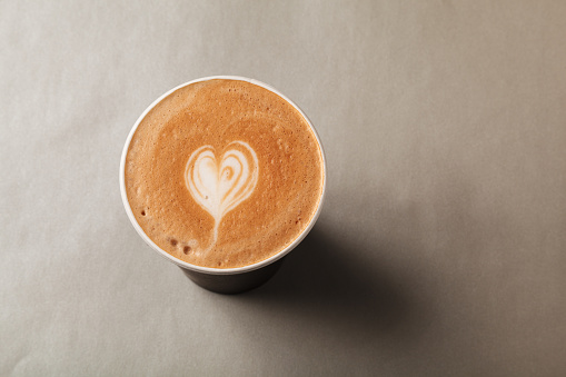 Craft cardboard cup of latte or cappuccino with patterned heart latte art stands on a grey background. Concept of serving coffee to take away, coffee to go