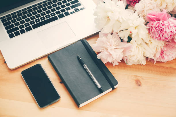 istock Stylish phone with empty screen, laptop, notebook, pen,  pink and white peonies on wooden table with space for text. Freelance concept. International womens day. Stylish working place. 1132563767