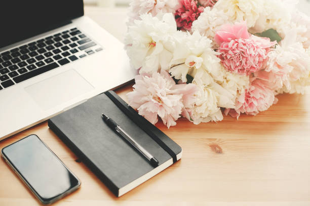 istock Stylish phone with empty screen, laptop, notebook, pen,  pink and white peonies on wooden table with space for text. Freelance concept. International womens day. Stylish working place. 1132565206