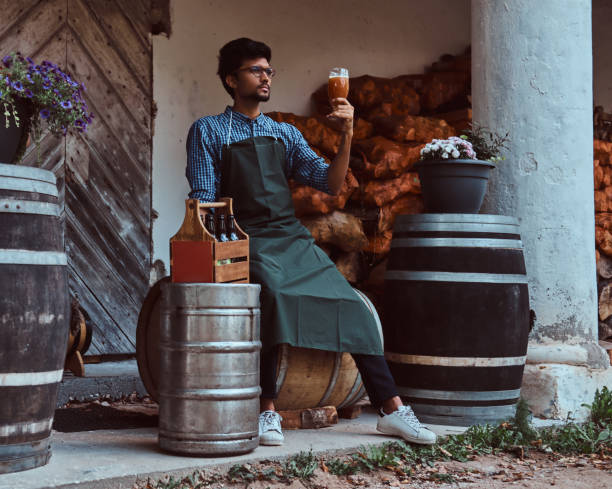 maître brasseur assis sur un tonneau en bois et tient un verre de bière artisanale, se détend après le travail. - after work beautiful people beer beer bottle photos et images de collection