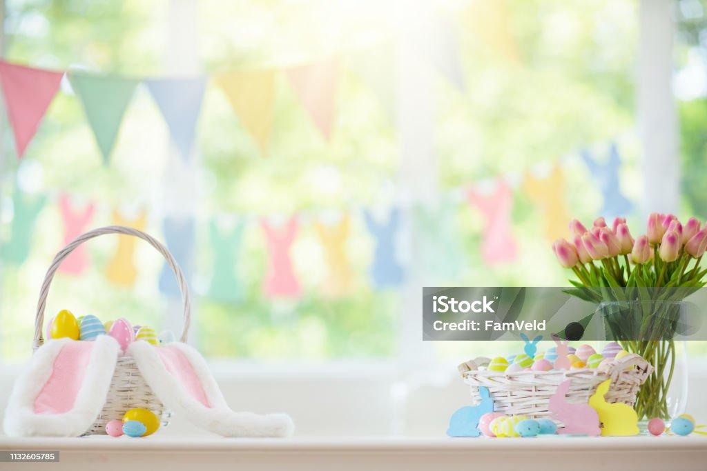 Easter home decoration. Eggs basket and bunny. Easter home decoration. Basket with colorful dyed eggs and rabbit ears, tulip flowers and wooden bunny for festive Easter celebration at home. Kids basket for Easter egg hunt. Easter Stock Photo