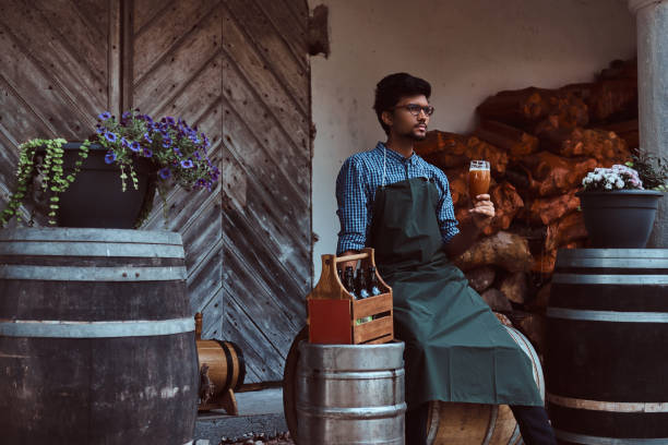 maître brasseur assis sur un tonneau en bois et tient un verre de bière artisanale, se détend après le travail. - after work beautiful people beer beer bottle photos et images de collection