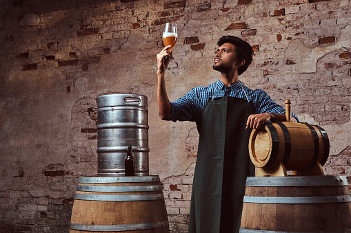 Expert brewer in apron standing near barrels checking quality of brewed drink at indi brewery factory.