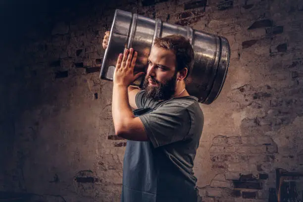 Bearded brewer in apron holds barrel with craft beer at brewery factory.