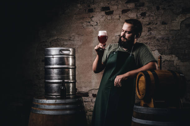 Brewer in apron standing near barrels and drinks craft beer at old brewery factory. stock photo
