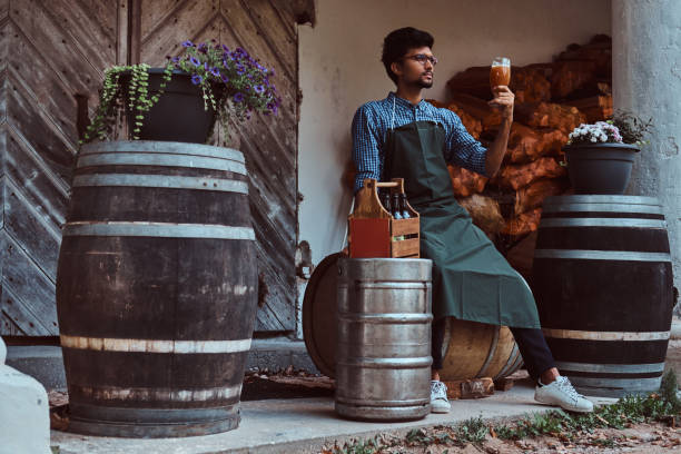 maître brasseur assis sur un tonneau en bois et tient un verre de bière artisanale, se détend après le travail. - after work beautiful people beer beer bottle photos et images de collection