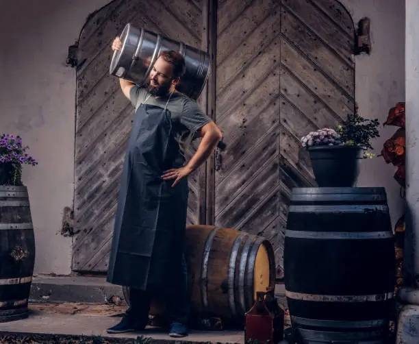 Bearded brewer in apron holds barrel with craft beer at brewery factory.