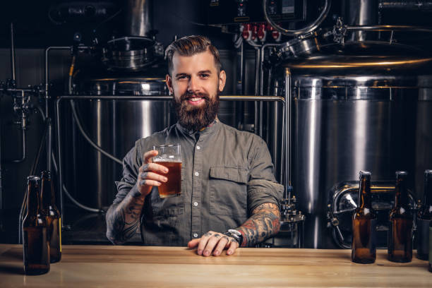 スタイリッシュな髭と髪を持つ入れ墨男性は、インディー醸造所のバーカウンターに座ってクラフトビールのパイントを保持しています。 - attack craft ストックフォトと画像