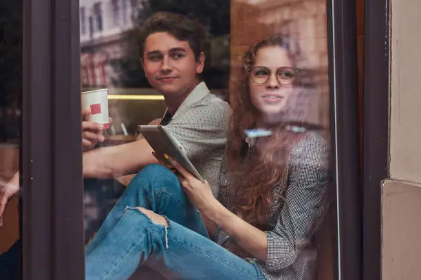 Happy young students drinking coffee and using a digital tablet sitting on a window sill at a college campus during a break.