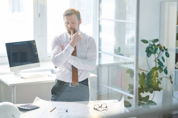 istock Serious pensive construction engineer in formalwear standing at table and keeping thumb on lips while thinking of solution for design project 1132617902