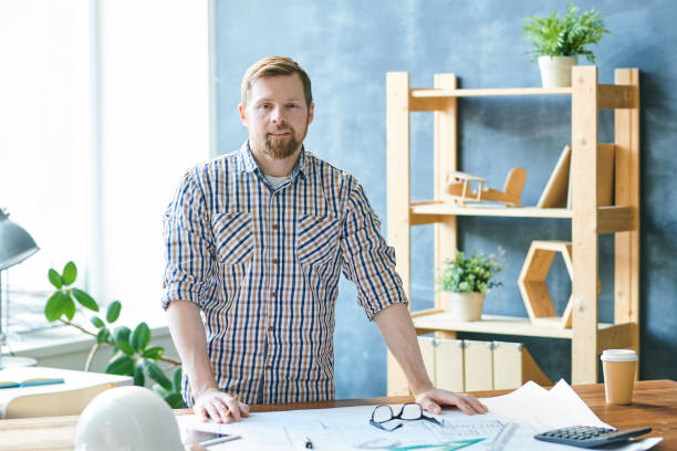 istock Content handsome young bearded engineer in casual shirt standing at desk and looking at camera while working with house project in office 1132618598