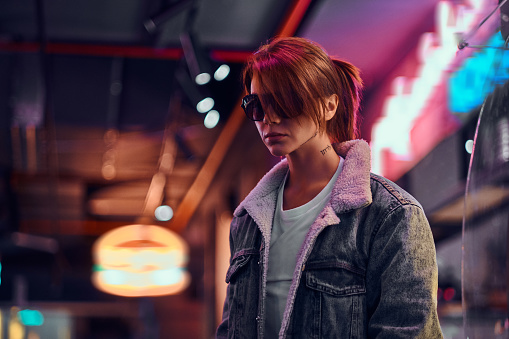 Stylish redhead girl with tattoo on her face wearing a denim coat and sunglasses standing in the night on the street. Shop signboards, neon, lights.