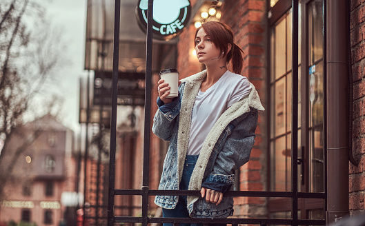 A beautiful tattooed girl wearing a denim coat holding cup with takeaway coffee outside near the cafe.