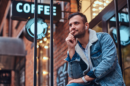 Portrait of a pensive stylish man wearing a denim jacket with wireless headphones holding takeaway coffee outside the cafe.