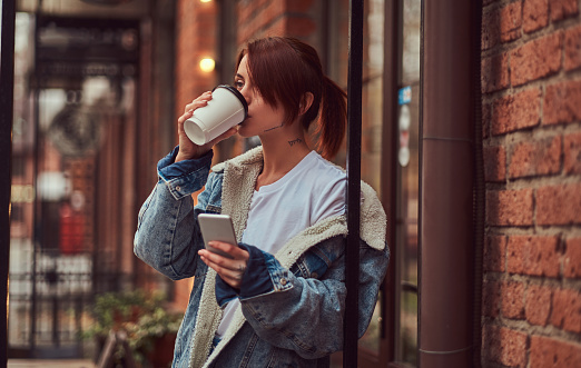 A beautiful girl wearing a denim coat drinks takeaway coffee holding a smartphone outside the cafe.