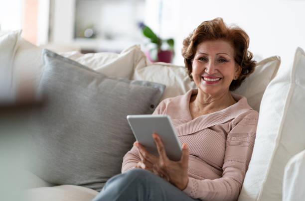 Beautiful senior woman facing camera smiling while holding tablet and relaxing on couch at home