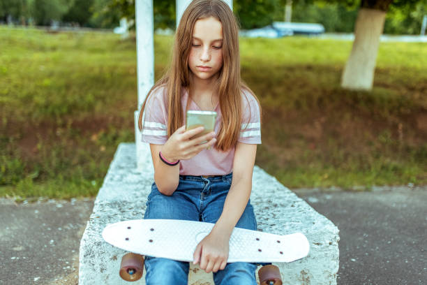 teen girl 12-13 years old, sitting in hands of a skate. in summer in city in casual jeans and pink t-shirt. communication in internet, in hands smartphone holds writes a message in application. - 12 13 years fotos imagens e fotografias de stock