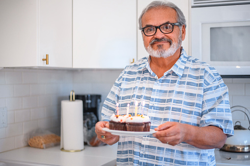 Happy senior man holding plate with candles on birthday cupcakes. Retired male is preparing for party in kitchen. He is wearing shirt and eyeglasses.