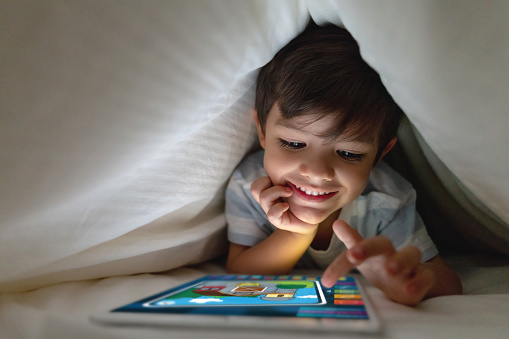 Portrait of a happy boy in bed playing games on a tablet computer and smiling under the covers