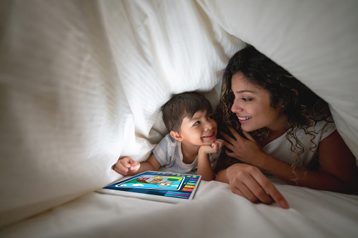 Portrait of a happy mother and son using a digital tablet to play in bed and smiling under the covers