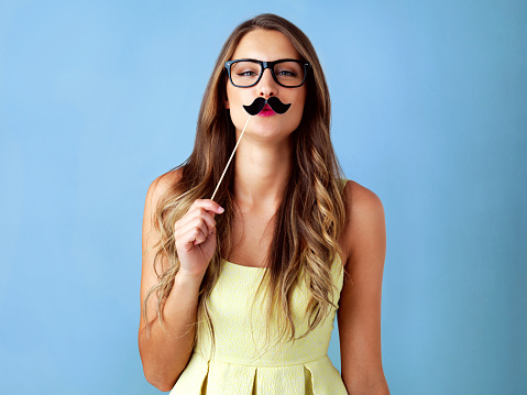 Studio shot of a young woman holding a moustache prop to her face against a blue background