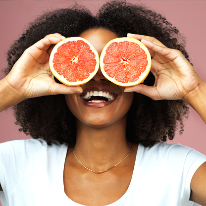 Studio shot of an attractive young woman covering her eyes with slices of grapefruit against a pink background