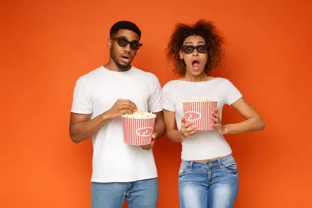 Young shocked african-american couple in 3d glasses watching scary movie, holding bucket of popcorn on orange background