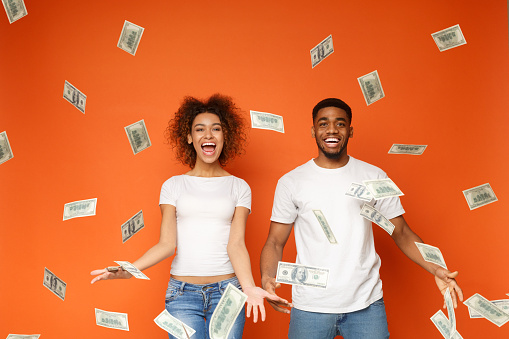 Financial success. Excited young african-american couple standing under money banknotes shower, orange background