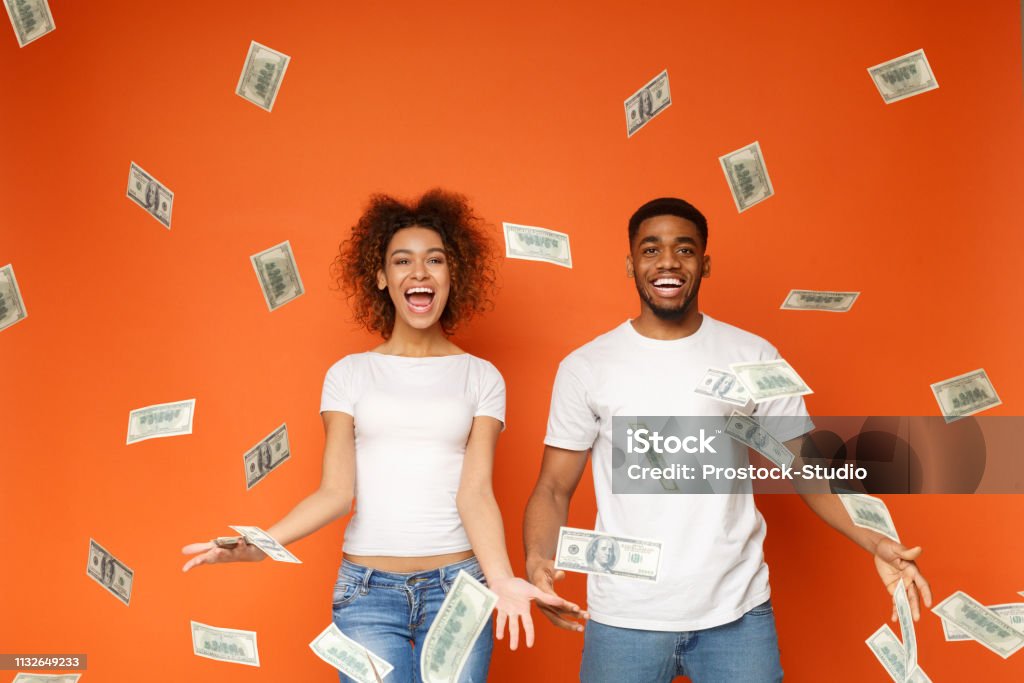 Young black couple standing under money banknotes shower Financial success. Excited young african-american couple standing under money banknotes shower, orange background Currency Stock Photo