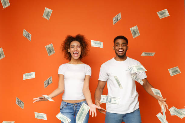 Young black couple standing under money banknotes shower Financial success. Excited young african-american couple standing under money banknotes shower, orange background money rain stock pictures, royalty-free photos & images