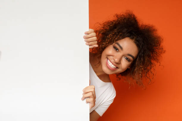 Black woman looking out of white blank card Smiling african-american woman looking out of white blank sign with copy space on orange background female likeness photos stock pictures, royalty-free photos & images