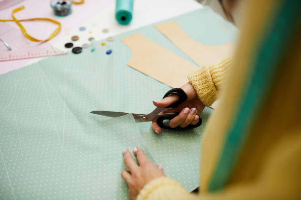 istock White seamstress in yellow jumper is cutting with big black scissors pattern contoured on light blue piece of cloth on table 1132658434